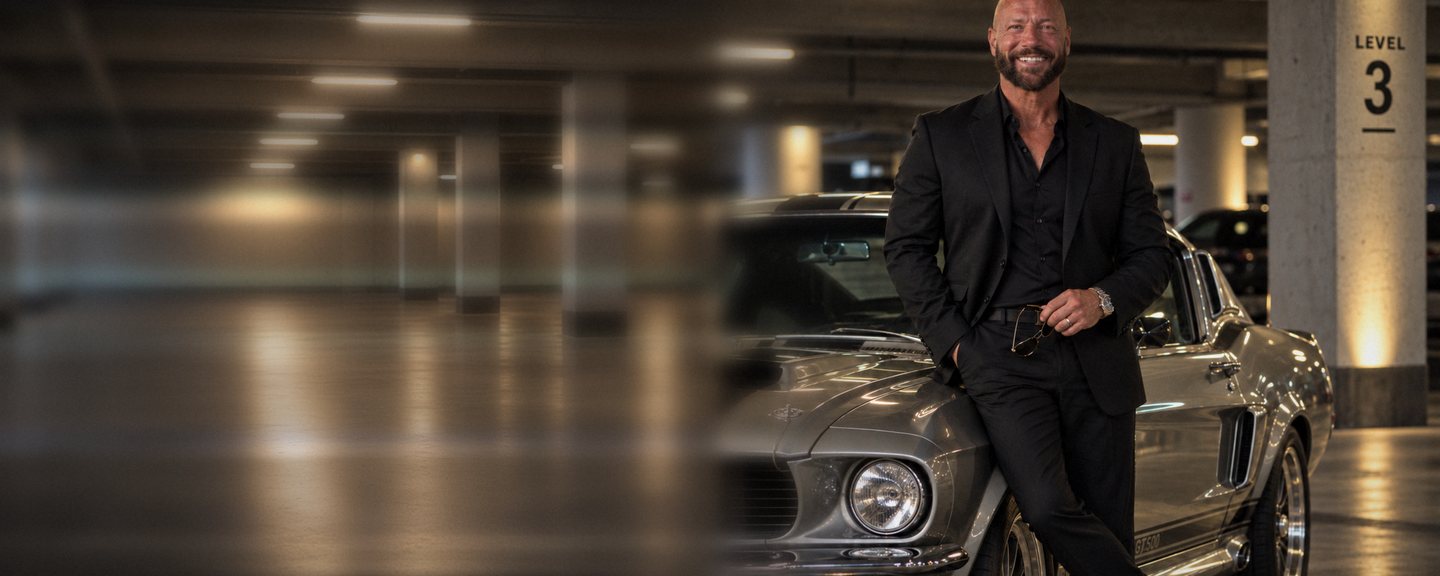 Chad Moore in a black jacket leaning on his silver 1967 Mustang fastback in a parking garage