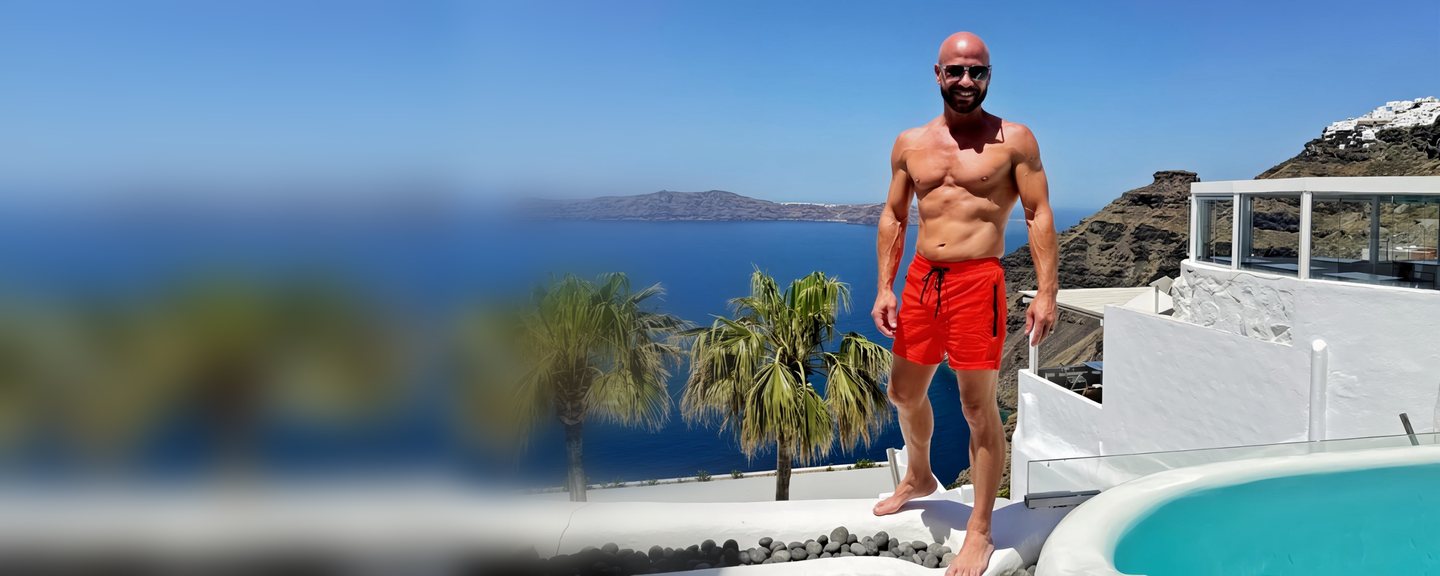 Chad Moore smiling on a whitewashed terrace in Santorini, Greece, above the blue Aegean Sea with palm trees and a pool