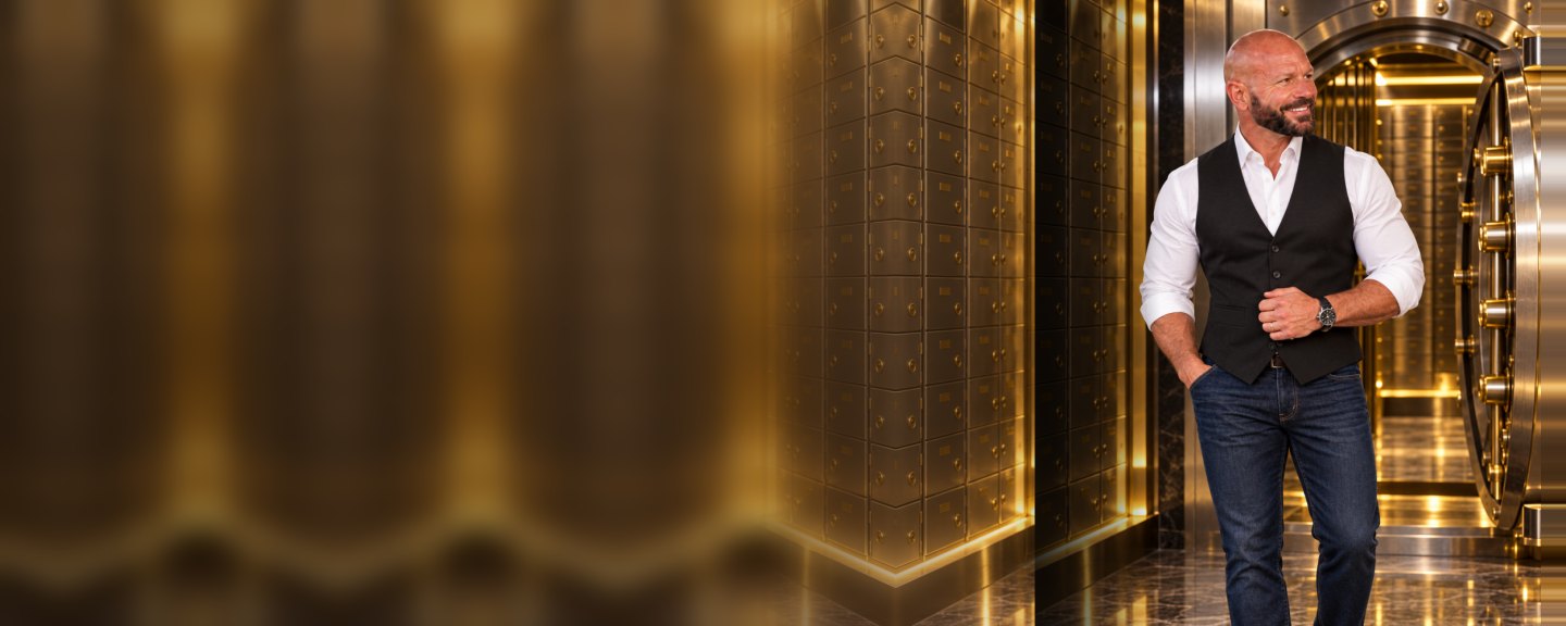 Chad Moore in a black vest, white dress shirt and jeans, smiling inside a gold-lit bank vault beside the open vault door